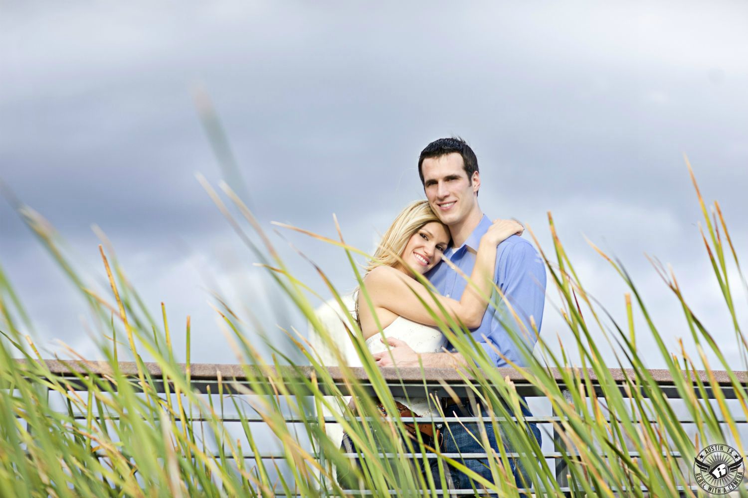 Happy blond bombshell wearing a white halter top, brown belt and dark blue jeans smiles and puts her head on the should of a happy, dark spiky haired guy wearing a blue button up dress shirt with the sleeve casually rolled up and blue jeans standing on a dock behind a railing and a lot of green reeds at Butler Park in this happy engagement photo in Austin, Texas. 
